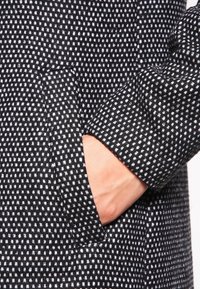 Cappotto a righe in bianco e nero con tessuto testurizzato, tasca laterale e design della manica aderente. Ingrandimento della mano nella tasca.
