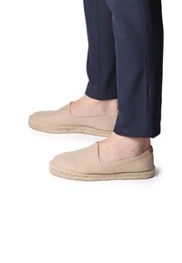 Beežid espadrillid, millel on kootud tekstuur ja elastne külg, jutana tald, esitatud valge tausta ees.