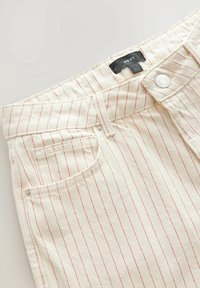 Pantalones de mezclilla beige con rayas verticales rojas. Cuenta con un cierre de botón estándar, pasadores para cinturón y un bolsillo frontal. Etiqueta de la marca visible.
