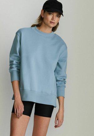Light blue sweatshirt with ribbed cuffs and hem. Worn over black cycling shorts. Model in a black cap with a neutral background.
