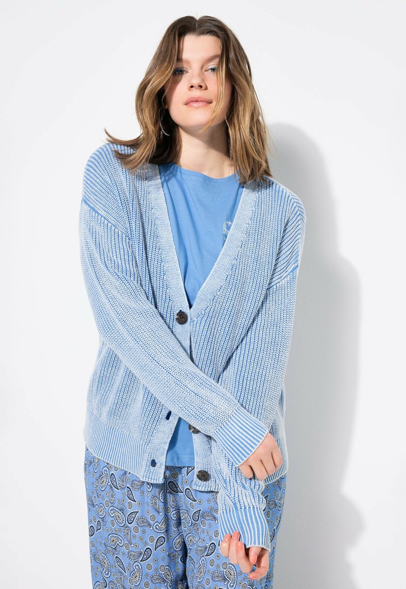 Vaalean sininen neulecardigan, jossa on V-pääntie, napit edessä ja ribbitekstuuri. Yhdistetty siniseen T-paitaan ja kuvioituihin pyjamahousuihin.