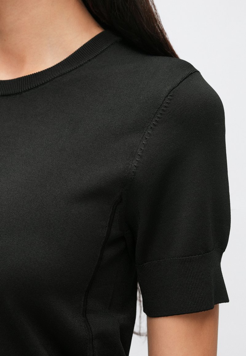 Sort kortærmet top lavet af ribstrikket stof. Har en rund halsudskæring og diskrete syningsdetaljer langs skuldersømmene.