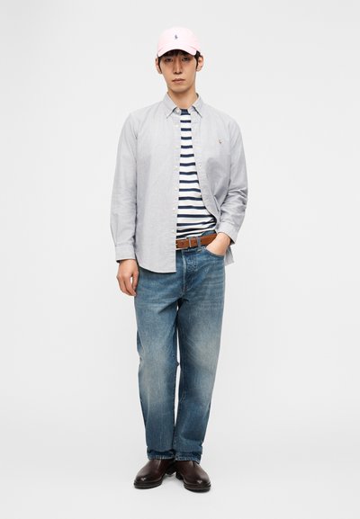 Νεαρός άνδρας που στέκεται φορώντας ένα ροζ καπέλο, ανοιχτό γκρι πουκάμισο με κουμπιά πάνω από μπλε μπλουζάκι με ρίγες, μπλε τζιν, καφέ ζώνη και σκούρα καφέ μπότες.