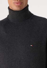 Maglione a collo alto grigio scuro in un tessuto a maglia morbida, con un piccolo logo rosso, bianco e blu sulla parte sinistra del petto.