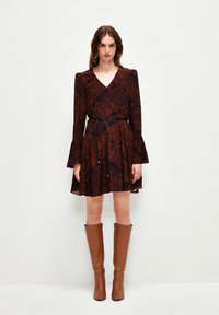 Wickelkleid in Dunkelrot mit schwarzem Blattmuster, langen Glockenärmel und taillierter Taille. Kombiniert mit knielangen braunen Stiefeln mit Absatz.