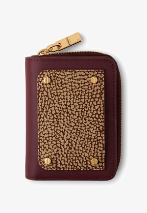 Portefeuille compact en cuir bordeaux avec un patch texturé beige fixé par quatre vis dorées et un zip doré gravé du nom de la marque.