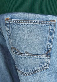 Jeans de mezclilla azul claro con un bolsillo trasero con costuras reforzadas, bordes deshilachados y una cintura acanalada verde en contraste arriba.