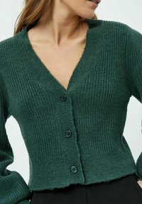 Cardigan verde a coste con scollatura a V, maniche lunghe e quattro bottoni neri sul davanti. L'orlo è irregolare per un aspetto testurizzato.