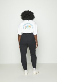 Henkilö, jolla on kiharat hiukset, seisoo kasvot seinää kohti valkoisessa kukkapaidassa, jossa on teksti "Always Original", mustissa housuissa ja valkoisissa lenkkareissa.