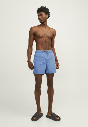 Shorts de bain bleu clair avec une taille élastique et un cordon de serrage. Portés avec des sandales à glissière noires. Modèle se tenant devant un fond neutre.