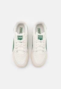 Paire de baskets blanches Puma avec des patchs verts du logo sur les languettes et des bandes latérales vertes, vues de dessus sur fond blanc.