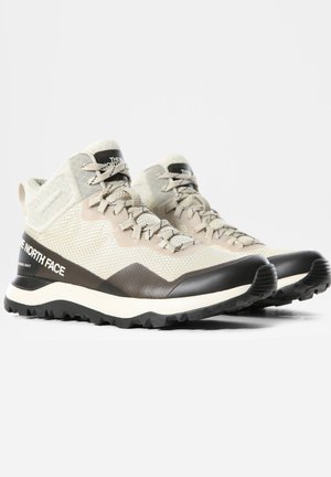 Zapatos de senderismo de caña media de color beige claro y negro con suelas robustas, parte superior de malla, con la marca "The North Face" en el lateral y la lengüeta, en ángulo para mostrar el frente y el lateral.