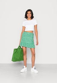 Saia verde com bolinhas brancas, bainha rendada, combinada com um top cropped branco; segurando uma mala de mão verde texturizada. Ténis brancos completam o visual.