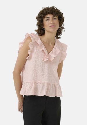 Haut texturé rose clair avec des manches à volants et un col en V, présentant un ourlet évasé. Porté avec un pantalon noir, mettant en valeur les détails du vêtement.
