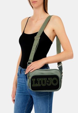 Donna che indossa una canottiera nera e jeans blu, con una borsa verde a tracolla con il logo "LIU JO" sulla tasca frontale.