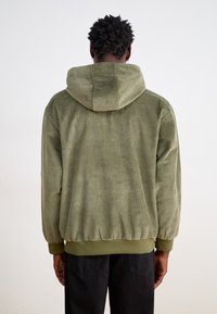 Olivgrön korddrickshuvtröja med dragsko i huvan, ribbade manschetter och nederkant; har vertikal ribbad textur. Sedd bakifrån.