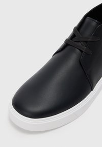 Sneaker din piele neagră, cu vârf rotunjit, șireturi gri și talpă de cauciuc albă. Textură netedă, cu detalii minime și un design elegant.