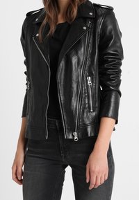 Kvinde iført en sort læder bikerjakke med sølv lynlåse over en sort top og mørk grå jeans, stående mod en hvid baggrund.