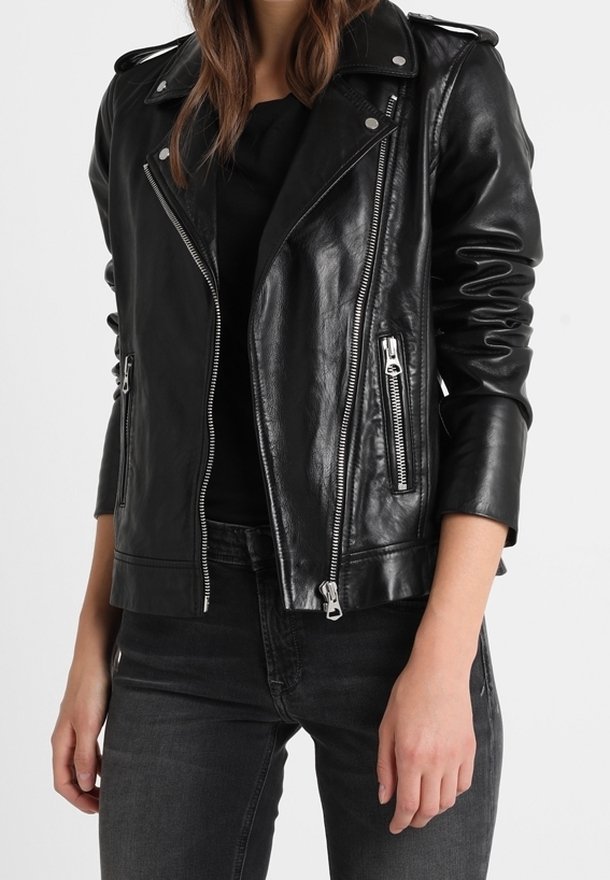 Kvinde iført en sort læder bikerjakke med sølv lynlåse over en sort top og mørk grå jeans, stående mod en hvid baggrund.