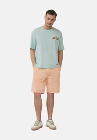 Jasnoniebieska bawełniana koszulka z pomarańczowym graficznym wzorem kwiatu, w połączeniu z brzoskwiniowymi szortami i beżowymi sneakersami z białymi akcentami.