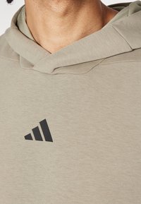Hoodie beige chiaro realizzato in morbido tessuto, con un grande logo Adidas nero sulla parte sinistra del petto. Dettagli in cucitura testurizzata sul cappuccio.