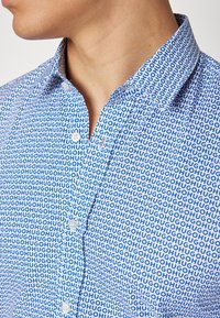 Camisa de manga corta fabricada con un tejido que presenta un patrón repetitivo en azul y blanco con la palabra "HUGO". Cuenta con un cuello y un cierre delantero de botones.