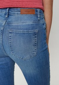 Zilā džinsu bikses ar gludu tekstūru, kurām ir viena aizmugurējā kabata un brūns ādas uzplecs ar uzrakstu "Nümph". Klasiska piegriezuma dizains.