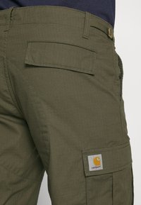 Pantalones cargo en verde oliva, hechos de una tela texturizada, con un bolsillo parche prominente y una etiqueta de logo cuadrada.