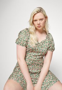 Vestido floral com mangas bufantes, apresentando uma base verde e pequenas flores rosas e brancas. Design ajustado com decote quadrado e comprimento curto.