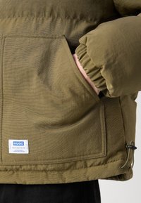 Main portant une manche de doudoune vert olive reposant dans une grande poche avant avec une étiquette de marque Hugo et un cordon de serrage réglable à l’ourlet.