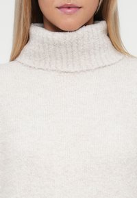 Maglione beige a collo alto in maglia, con colletto a coste e una finitura morbida e testurizzata, caratterizzato da una vestibilità rilassata e un design minimalista.
