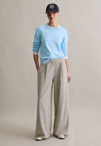 Lichtblauwe trui met lange mouwen, gecombineerd met beige en zwart geruite wijde broekspijpen en een zwarte pet met gouden cijfers.