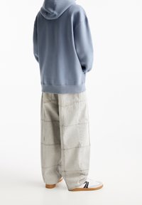 Persona con un hoodie azul, pantalones sueltos de parcheo en gris claro y zapatillas blancas con rayas negras y suelas de goma, de pie sobre un fondo blanco.