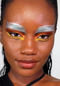 Close-up van een persoon met gedurfde make-up bestaande uit zilver en oranje oogschaduw, gele accenten en glanzende lippen. De huid heeft een zachte textuur.