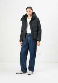 Jeune femme portant une doudoune noire, un t-shirt blanc, un jean bleu à jambes larges, des chaussures blanches, tenant un petit sac à main blanc devant un fond uni.