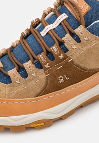 Bruna mocka- och blå denimskor med intrikat sömnad, gummisula och gul Vibram-logotyp. Har snörning och mjuka texturer.