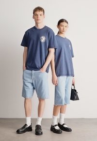 Tamsiai mėlyni grafikos marškinėliai derinami su šviesiais džinsiniais šortais. Vyras modelis dėvi juodus loaferius, o moteris modelė neša mažą juodą rankinę.