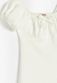 Weißes, strukturiertes Oberteil mit kurzen Puffärmeln, einem runden Ausschnitt und einem vorderen Bindedetail. Der Stoff hat eine leicht zerknitterte Optik.