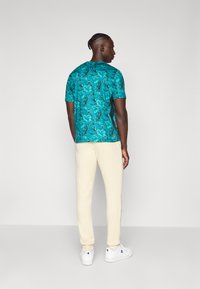 Man met gevlochten haar die een turquoise gestreept shirt met korte mouwen, crèmekleurige joggingbroek en witte sneakers draagt, staand voor een effen grijze achtergrond.