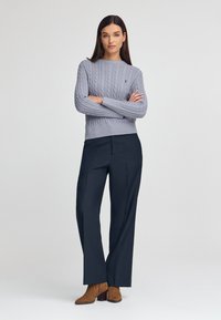 Jasnoszary sweter z dzianiny w warkocze z prążkowanymi mankietami, zestawiony z granatowymi spodniami o wysokim stanie i brązowymi zamszowymi botkami sięgającymi kostek.