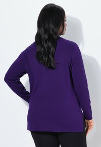Vrouw met lang zwart haar, gekleed in een paarse trui met lange mouwen en zwarte broek, die met haar rug naar de camera staat tegen een witte achtergrond.