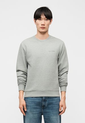 Νέος άνδρας με ίσια μαύρα μαλλιά φοράει ανοιχτό γκρι μακρυμάνικο φούτερ και μπλε τζιν, στέκεται μπροστά σε απλό λευκό φόντο.