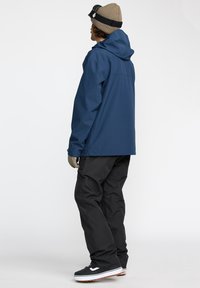 Veste imperméable bleu marine avec capuche, associée à un pantalon noir et des chaussures noires avec des semelles blanches. Présente une coupe décontractée et un matériau durable.