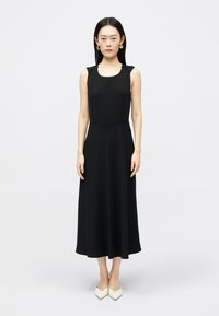 Γυναίκα που στέκεται σε ένα αμάνικο μαύρο μίντι φόρεμα, φορώντας χρυσά σκουλαρίκια λουλούδια και λευκές μυτερές γόβες, σε φόντο λευκό.