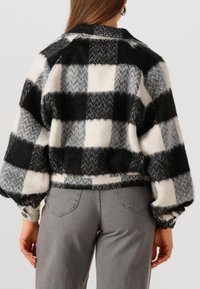 Vrouw die een zwart-wit geblokte fuzzy jas met opgezette mouwen en grijze jeans draagt, gefotografeerd van achteren tegen een witte achtergrond.