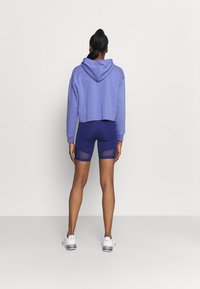 Felpa viola tagliata con tasca a marsupio frontale, abbinata a pantaloni corti blu navy con accenti in rete. Il modello indossa scarpe da ginnastica bianche.