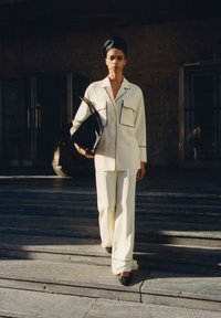 Ivory outfit met een button-down shirt met zwarte piping en twee voorkeiten, gecombineerd met losse broek en zwarte hakken. Het model houdt een zwarte tas vast.