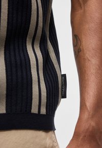 Κοντινό πλάνο ενός ατόμου που φοράει πλεκτό πάνω μέρος με μαύρες και μπεζ ρίγες, με ετικέτα της μάρκας Marc O'Polo στο πλάι, και τατουάζ στο αντιβράχιο.