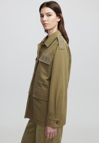 Chaqueta utilitaria caqui con cierre de botones, parches en los hombros y múltiples bolsillos frontales. Presenta una tela texturizada y un corte relajado.