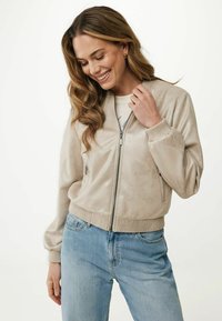 Beżowa kurtka bombera z miękkiego, błyszczącego materiału, z przednim zamkiem, ściągaczami na mankietach i u dołu. Noszona z jasnoniebieskimi dżinsami.